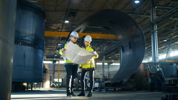 男性和女性工业工程师查看项目蓝图 同时站在大型重工业制造工厂中间的管道部件周围 — 图库照片