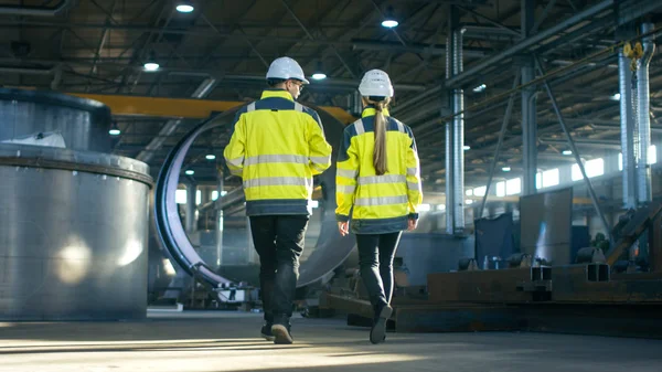 后视拍摄的男性和女性工业工程师在步行通过重工业制造厂的讨论 大型金属制品结构 管线元素围绕 — 图库照片