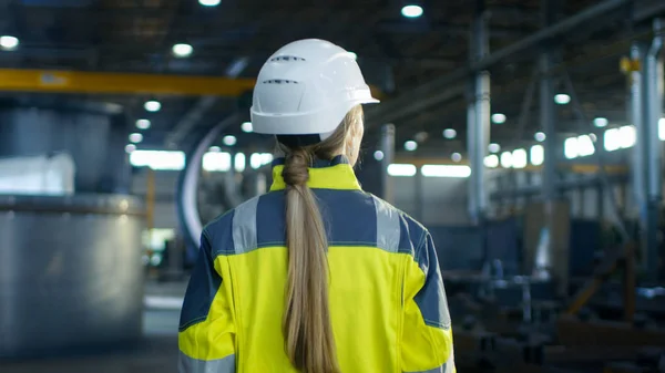 Shot of Female Industrial Worker in the Hard Hat Walking Through Heavy Industry Manufacturing Factory. In the Background Various Metalwork Project Parts Lying