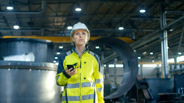 在硬帽中的女工业工人在穿过重工业制造厂时使用手机 在背景下各种金属制品工程零件的谎言 — 图库照片