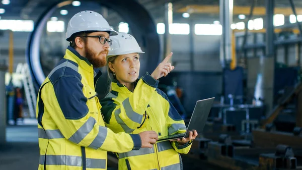 在硬帽子的男性和女性工业工程师的肖像讨论新的项目 同时使用笔记本电脑 他们戴着安全夹克 在重工业制造工厂工作 — 图库照片