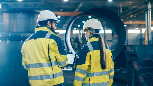 男性和女性的工业工程师使用笔记本电脑 并在走过重工业制造厂时进行讨论 他们穿着安全帽和安全夹克 在背景大金属制品结构 管道组件 — 图库照片