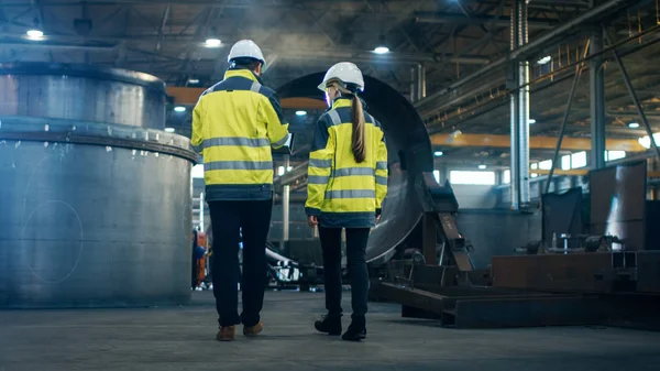 男性和女性的工业工程师使用笔记本电脑 并在走过重工业制造厂时进行讨论 他们穿着安全帽和安全夹克 — 图库照片