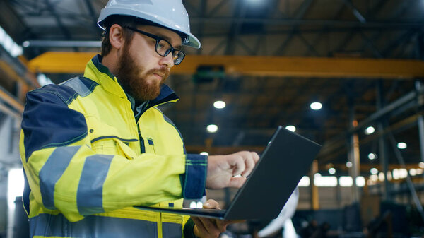 Промышленный инженер в твердой шляпе носить пиджак безопасности использует сенсорный экран ноутбука. Работает на заводе тяжелой промышленности. На заднем плане находятся процессы сварки / металлообработки
.