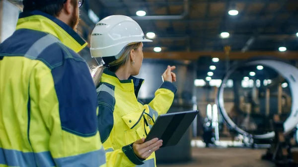 在安全帽和安全夹克的男性和女性工业工程师在使用平板电脑时讨论新项目 他们在重工业制造厂工作 — 图库照片