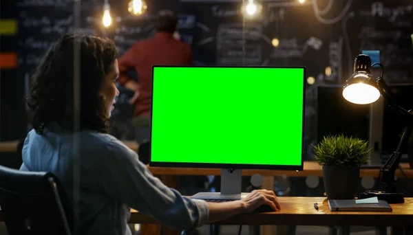 美丽的女性办公室员工在她的桌面工作在一个模拟的绿色屏幕个人计算机 在肩膀上的镜头 她的同事在后台工作 创意办公室晚上 — 图库照片