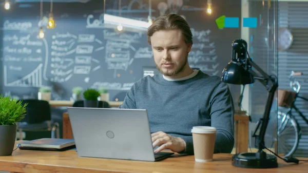 Handsome Young Office Employee Thinks on a Problem Solution While Typing on a Laptop Computer. He\'s Working in the Creative Stylish Office.