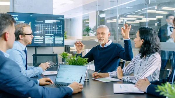 Senior Executive Explains Company\'s Vision and Potential to His Employees. They are Sitting at Big Table in Meeting Room. TV Screen on the Wall Shows Corporate Growth.