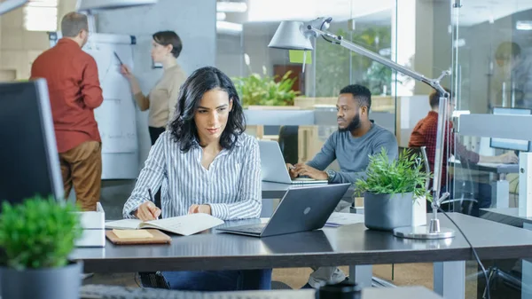 Busy International Office, Beautiful Hispanic Woman Working at Her Desk on a Laptop, She Writes Down information in Her Notebook. In the Background Her Coworkers.