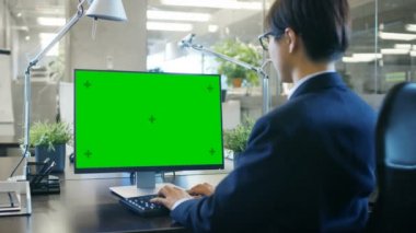 In the Office Businessman works at His Desk on a Personal Computer with Mock-up Green Screen. Colleague Enters Office and Takes Place at Her Desk.