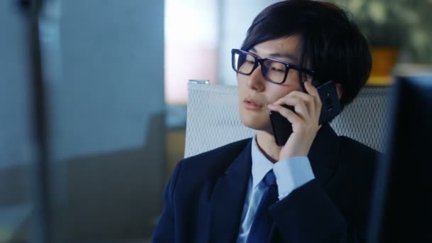 日本商人穿着西装和眼镜 坐在办公桌前 在手机上交谈 在个人电脑上工作的肖像 — 图库视频影像