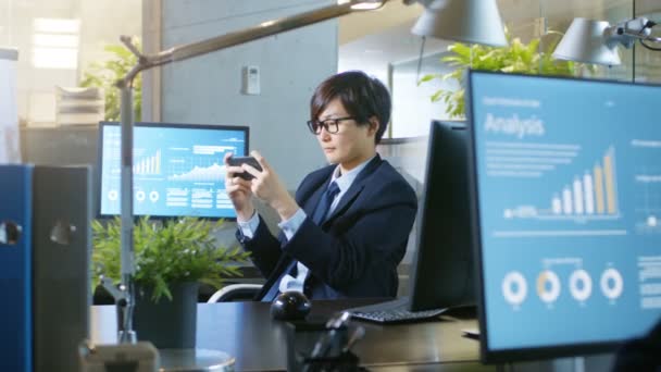彼のスマート フォン上東アジアの実業家 Plaing ビデオゲームのドリー ズーム ショット スタイリッシュなオフィスで自分の机に座っている笑顔のハンサムな男 — ストック動画