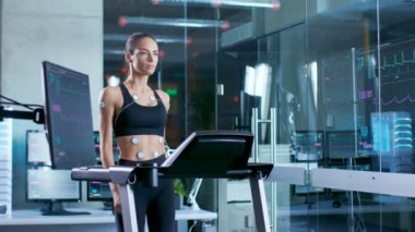 Ona bağlı elektrotlar ile spor sutyen giyen güzel bir kadın atlet spor bilim laboratuarında bir koşu bandı üzerinde yürür. Monitörler Ekg gösterilen veri ile arka plan laboratuvarda.