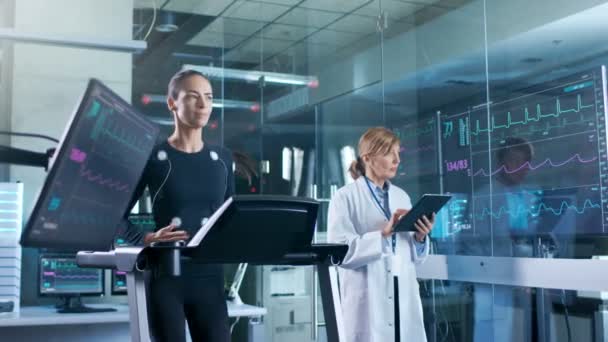 女运动员在跑步机上运行 电极贴在她的身体 而科学家持有平板电脑监督整个过程 在背景实验室与显示器显示 Ekg — 图库视频影像