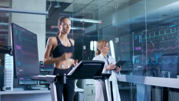 美丽的女运动员运行在跑步机上与电极连接到她的身体 女医生使用平板电脑和控制 Ekg 数据显示在实验室监视器 — 图库视频影像