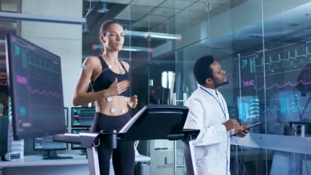 Beautiful Woman Athlete Runs on a Treadmill with Electrodes Attached to Her Body, Physician Uses Tablet Computer and Controls EKG Data Showing on Laboratory Monitors. — Stock Footage Beautiful Woman Athlete Runs Treadmill Electrodes Attached Her Body Physician Video Clip