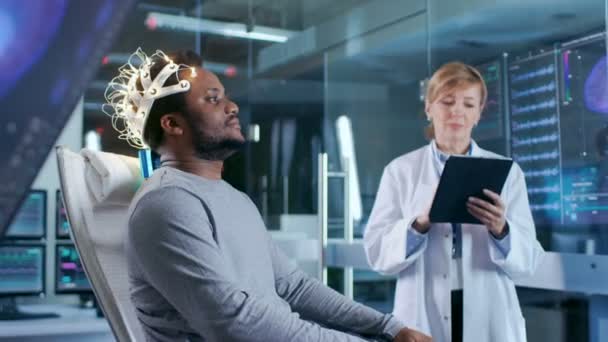男子穿着脑波扫描耳机坐在椅子上 而科学家与平板电脑监督过程 现代脑研究中的实验室监测显示英语阅读和脑模型 — 图库视频影像