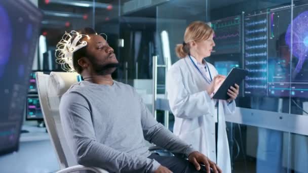 男子穿着脑波扫描耳机坐在椅子上 而科学家与平板电脑监督过程 现代脑研究中的实验室监测显示英语阅读和脑模型 — 图库视频影像