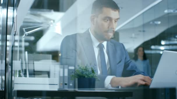 自信を持ってハンサムな実業家は 彼の机でノート パソコンで動作します ガラス張りのモダンなオフィスにスタイリッシュな男 — ストック動画