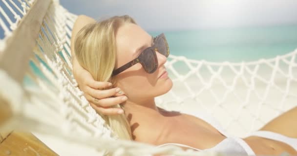 田园诗般的场景 特写镜头的一个美丽的金发碧眼的女人在太阳镜日光浴躺在海滩上的吊床 Azure 海滩与白沙和海蓝宝石水 — 图库视频影像