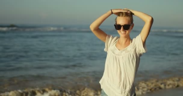 美丽的金发女郎沿着海滩散步 她在跳舞和呼啸 举起她的手臂和微笑 背景中的海洋与自然 — 图库视频影像