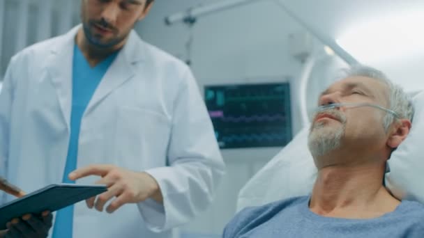 シニア男のベッドで横になっている病院でタブレット コンピューターを使った彼を診断した医師と看護師との協議します 技術により 近代的な病院病棟治療 — ストック動画