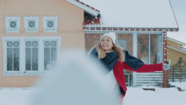 美丽的年轻微笑的女人跳和旋转下降雪 身穿红衣的女孩 穿着围巾 在院子里享受冬日 站在背景中的田园诗之家 — 图库视频影像