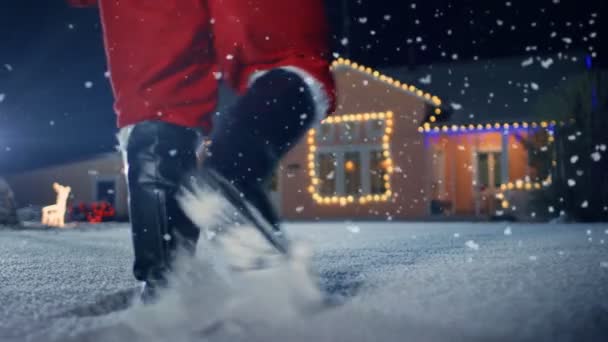 肩に赤いバッグを運ぶサンタ クロースは ライト 花輪で飾られた牧歌的な家の前庭に歩きます サンタをもたらす贈り物とプレゼント 魔法の大晦日の夜 低角度のスローモーション映像を次 — ストック動画