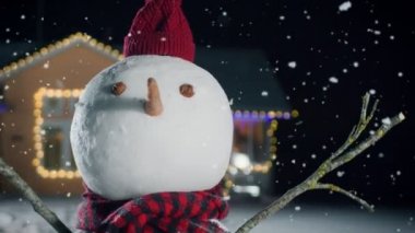 Komik kardan adam üzerinde yakın çekim giyen şapka ve atkı pastoral evin arka bahçesinde duran çelenk ile Noel arifesinde dekore edilmiştir. O büyülü kış akşam kar düşüyor.