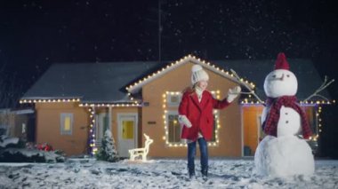 Sevimli küçük kız ishal evden aracılığıyla ön bahçede. Çocuk evin arka planda kış akşamında eğlenmek çelenk süslenmiş. Noel ağacı ve bahçede duran kardan adam.