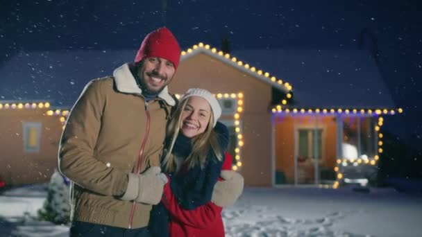 美しいカップルを愛情を込めて雪で受け入れます その牧歌的な家で飾られた花輪の背景の庭 一緒に若い家族楽しむ冬夜 — ストック動画