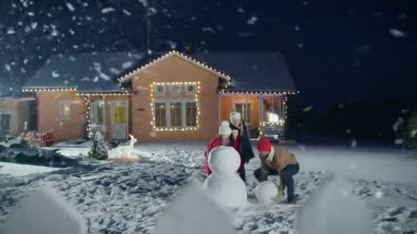 glückliche junge Familie beim Schneemannbasteln im Hinterhof ihres idyllischen Hauses. Vater rollt Schneeball, Tochter und Frau helfen ihm. Familie verbringt einen Winterabend zusammen.