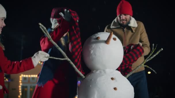 幸せな若い家族の作る雪だるまはそれ彼らの牧歌的な家の裏庭には 積雪です かわいい小さな娘と妻をスカーフで雪だるまをラップします クリスマスイブに一緒に時間を過ごす家族 — ストック動画