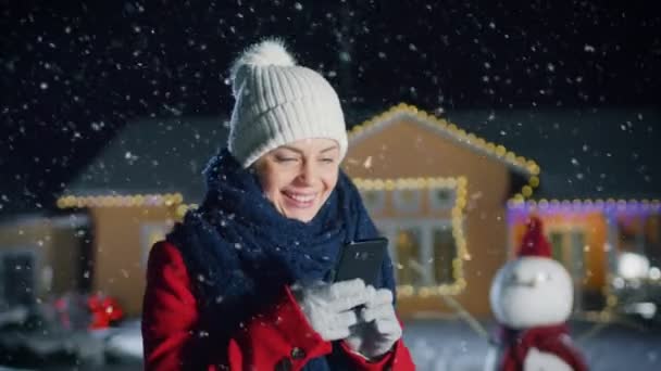 美しい若い女性を使用してスマート フォン クリスマスイブに裏庭の彼女牧歌的な家に飾られている花輪と一緒に立っています スカーフと赤い上着 彼女の家族や友人の携帯電話を使用するハッピーなクリスマス メッセージが送信の女の子 — ストック動画