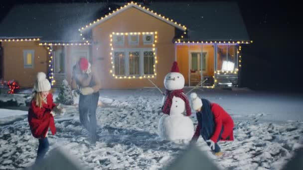 幸せな若い父 母とかわいい小さな娘が雪玉で遊ぶ 花輪で飾られた牧歌的な家の裏庭で走り回ってしまって雪だるまを構築します 家族を費やしている時間一緒に つの冬の夜に 雪が降る — ストック動画