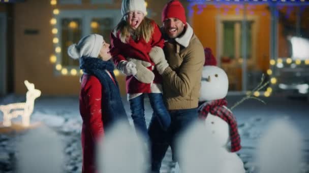 快乐的年轻家庭肖像在落雪 父亲举行女儿和拥抱他的妻子 微笑的家庭享受冬季假期在他们的田园诗般的房子装饰与花环的前院 — 图库视频影像