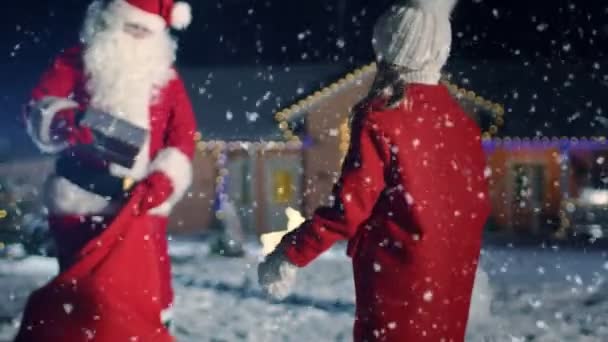 本格的なサンタ クロースは赤袋ラップ ギフト ボックスと与えるそれに出てかわいい女の子 バック グラウンドで牧歌的な装飾が施された前庭 雪だるま クリスマス ツリー 牧歌的な家 — ストック動画