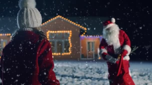 正宗的圣诞老人给可爱的小女孩一个包裹的礼品盒 在背景音乐装饰的前院 与雪人 圣诞树和田园诗般的房子 神奇的新年之夜与降雪 — 图库视频影像