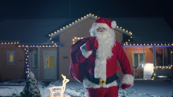 本格的なサンタ クロースを運ぶ赤バッグ肩 ライト 花輪で飾られた牧歌的な家の散歩 サンタをもたらす贈り物とプレゼント 雪が降ると魔法の大晦日の夜 — ストック動画