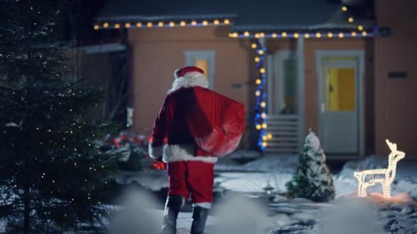 Der echte Weihnachtsmann, der eine rote Tasche über der Schulter trägt, betritt den Vorgarten des idyllischen Hauses, das mit Lichtern und Girlanden geschmückt ist. Weihnachtsmann bringt Geschenke und Geschenke. Zauberhafter Silvesterabend mit Schneefall.