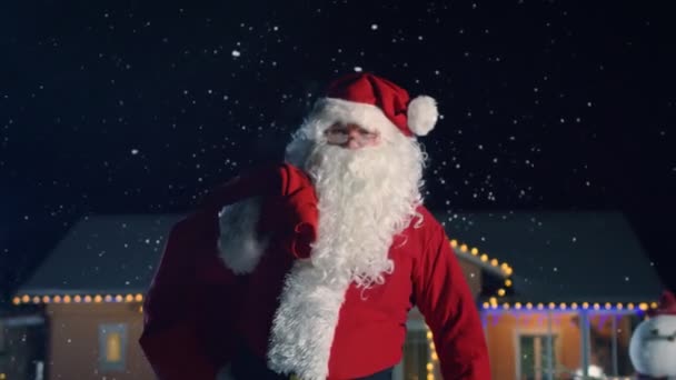 Autentické Santa Claus červená brašna na přes rameno, chodí ven z idylické dům vyzdoben světly a girlandy. Santa přináší dary a dárky. Kouzelný Silvestr s padající sníh.