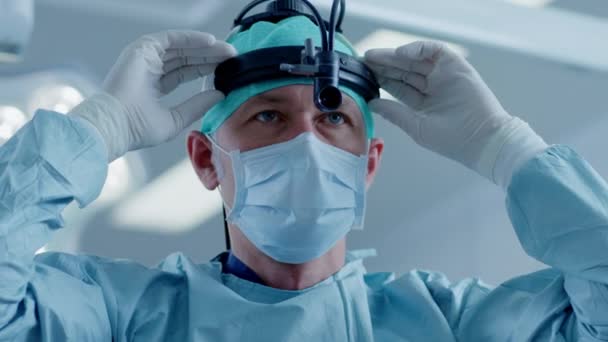 Na sala de operações do hospital, o cirurgião profissional coloca a lanterna cirúrgica, liga-a e olha para a câmera . — Vídeo de Stock