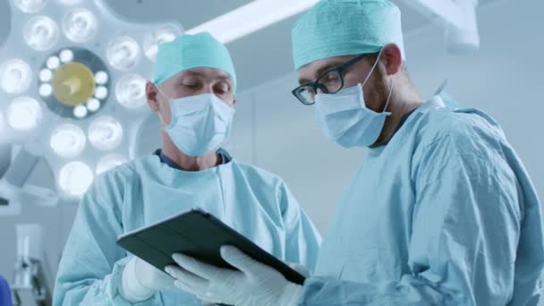 Два профессиональных хирурга используют цифровой планшетный компьютер, стоя в операционной современной больницы. — стоковое видео