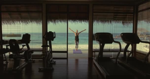 Back View shot van een fit jonge vrouw doet yoga op een veranda van een gym hut. Op de achtergrond exotisch eiland met prachtige kalme zee en heldere zonnige hemel. — Stockvideo