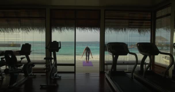Back View Shot av en fit ung kvinna gör yoga på en veranda i ett gym Hut. I bakgrunden exotiska ön med vackra lugna havet och klar solig himmel. — Stockvideo