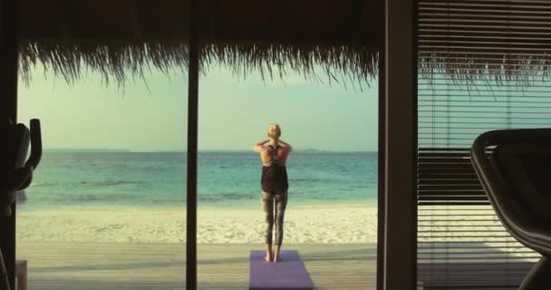 Back View shot van een fit jonge vrouw doet yoga op een veranda van een gym hut. Op de achtergrond exotisch eiland met prachtige kalme zee en heldere zonnige hemel. — Stockvideo