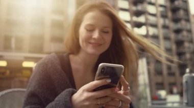Güzel Kızıl Saçlı Kadın Bir Bankta Otururken Akıllı Telefon Kullanır Gülümseyerek, Bağlı Kızlar Mesajlar Yazıyor. Arka Planda Muhteşem New York Cirty Cityscape.