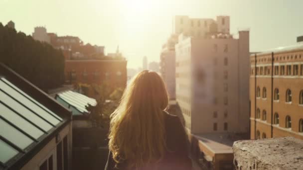 Back View shot van mooie vrouw staande op een dak, wind Shuffles haar lange rode haren. Stedelijk stadsgezicht uitzicht op New York City met grote wolkenkrabbers en gebouwen. — Stockvideo