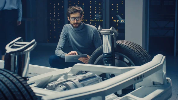 Ingénieur de conception automobile assis à côté du prototype de plate-forme de châssis de voiture électrique hybride, utilisant un ordinateur tablette pour l'amélioration de la conception. Installation avec châssis de véhicule avec suspension, roues, moteur et batterie — Photo