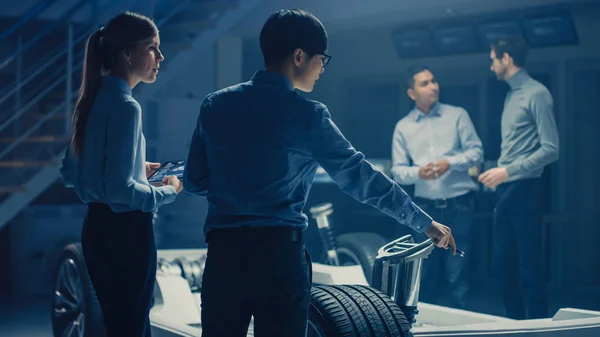 Ingenieros automovilísticos discuten y diseñan la plataforma eléctrica del chasis del coche, usando las computadoras de la tableta con el software 3D CAD. En el marco del vehículo de las instalaciones de innovación automotriz con ruedas, motor y batería — Foto de Stock